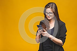 Portrait of smiling business woman in eyeglasses holding smartphone and looking back over yellow background. Using phone