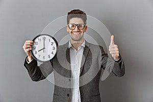 portrait of smiling businessman with clock in hand showing thumb up on grey