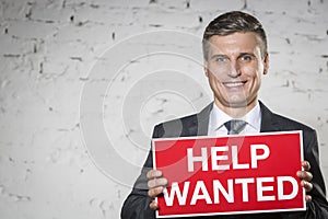 Portrait of smiling businessman holding help wanted sign while standing against white brick wall