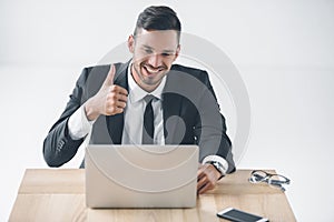 portrait of smiling businessman showing thumb up at workplace with laptop