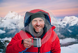 Portrait of smiling at camera high altitude mountaineer dressed red warm dawn jacket holding metal mug of hot tea in with