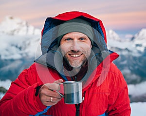 Portrait of smiling at camera high altitude mountaineer dressed red warm dawn jacket holding metal mug of hot tea in with