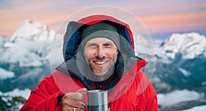 Portrait of smiling at camera high altitude mountaineer dressed red warm dawn jacket holding metal mug of hot tea in with