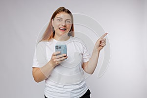 Portrait of a smiling casual woman holding smartphone and pointing finger away at copy space over white background. 30s