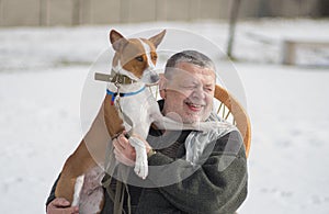 Portrait of smiling Caucasian senior man taking his cute basenji dog on the hands while sitting in a wicker chair