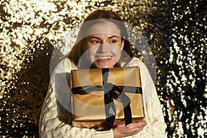 Portrait of a smiling caucasian woman holding a golden gift with a black ribbon on a golden sequin background