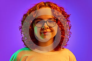 Portrait of smiling, cheerful teen girl with curly redhead hair, in glasses, lookin at camera against purple background