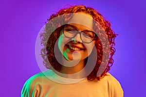 Portrait of smiling, cheerful teen girl with curly redhead hair, in glasses, lookin at camera against purple background