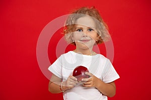 Portrait of smiling cherubic blue-eyed little girl with short curly fair hair holding big red apple on red background.