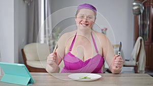 Portrait of smiling confident fat woman sitting at table with plate of cucumber slices. Motivated Caucasian obese lady