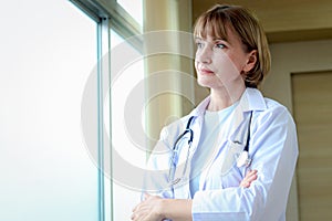 Portrait of smiling confident female doctor in white workwear with stethoscope, standing with arms crossed and looking through the