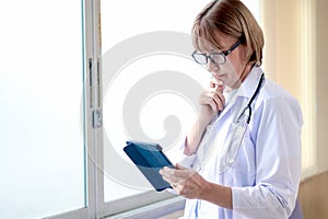 Portrait of smiling confident female doctor in workwear with stethoscope wearing glasses, holding tablet and standing by window