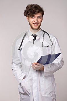 Portrait of smiling confident young smiling medical doctor with sthetoscope and clipboard over light gray background