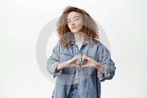 Portrait of smiling curly girl, stylish girlfriend shows heart, love sign and pucker lips, kissing gesture, standing in