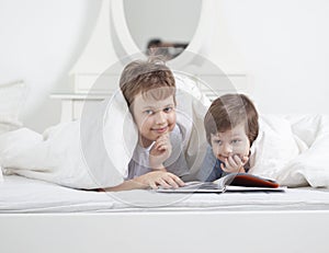 Portrait of a smiling cute brother reading a book lying and relax in bed in a bright white room