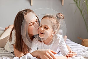 Portrait of smiling cute little girl lying in bed with mother, woman kissing her daughter with love