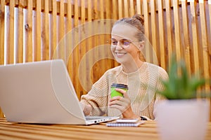 Portrait of smiling delighted satisfied woman freelancer with bun hairstyle wearing beige sweater working on laptop and drinking