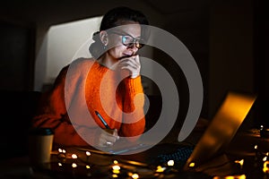 Portrait of smiling designer girl, working at graphic tablet on laptop. Wearing eyeglasses and orange sweater. In dark room home.