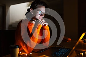 Portrait of smiling designer girl, working at graphic tablet on laptop. Wearing eyeglasses and orange sweater. In dark room home.