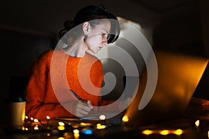 Portrait of smiling designer girl, working at graphic tablet on laptop. Wearing eyeglasses and orange sweater. In dark room home.