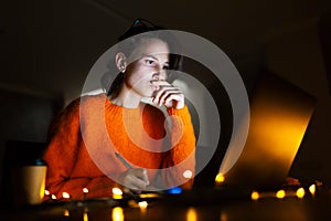 Portrait of smiling designer girl, working at graphic tablet on laptop. Wearing eyeglasses and orange sweater. In dark room home.