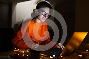 Portrait of smiling designer girl, working at graphic tablet on laptop. Wearing eyeglasses and orange sweater. In dark room home.