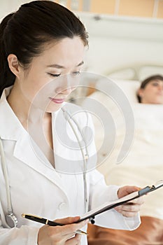 Portrait of smiling doctor holding a medical chart with patient lying in a hospital bed in the background