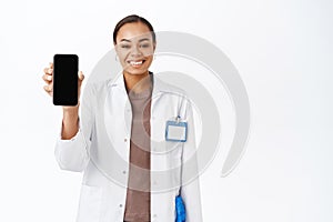 Portrait of smiling doctor shows her mobile phone screen, recommends online medical consultation app, standing over