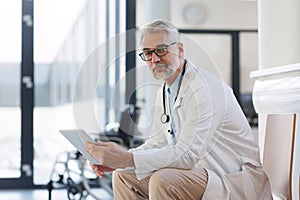 Portrait of smiling doctor sitting in hospital corridor. Handsome doctor with gray hair wearing white coat, stethoscope