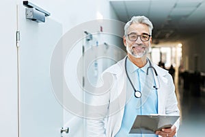 Portrait of smiling doctor standing in hospital corridor. Handsome doctor with gray hair wearing white coat, stethoscope