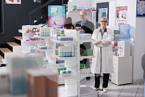Portrait of smiling elderly pharmacist looking at camera while standing with arm crossed in drugstore