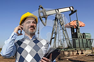 Portrait of Smiling Engineer With Mobile Phone In Front Of Petroleum Industry Oil Well Pump Jack