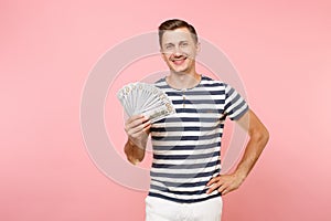 Portrait of smiling excited young man in striped t-shirt holding bundle lots of dollars, cash money, ardor gesture on
