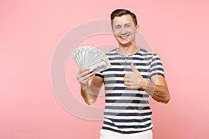 Portrait of smiling excited young man in striped t-shirt holding bundle lots of dollars, cash money, ardor gesture on