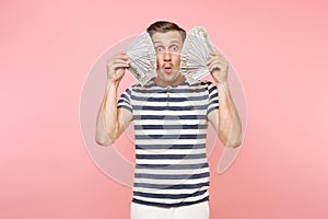 Portrait of smiling excited young man in striped t-shirt holding bundle lots of dollars, cash money, ardor gesture on