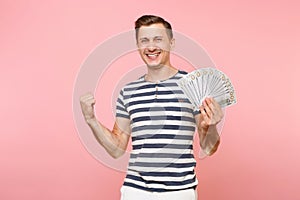 Portrait of smiling excited young man in striped t-shirt holding bundle lots of dollars, cash money, ardor gesture on