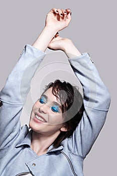 Portrait of smiling female in blue jacket with hands up. Woman with unusual beauty makeup and wet hair, and blue shadows make-up