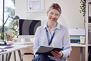 Portrait Of Smiling Female Doctor Or GP In Office With Clipboard