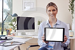 Portrait Of Smiling Female Doctor Or GP Sitting At Desk In Office Holding Digital Tablet