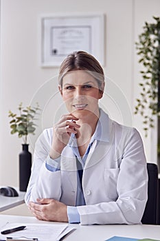 Portrait Of Smiling Female Doctor Or GP Wearing White Coat In Office Sitting At Desk