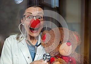Portrait of a smiling female doctor with red clown nose holding teddy bear, looking at camera.