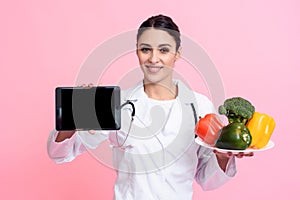 Portrait of smiling female doctor with stethoscope holding plate of vegetables and tablet isolated.