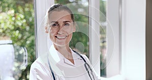 portrait of smiling female doctor with stethoscope standing by the window in hospital hallway. looking at camera. medical staff,