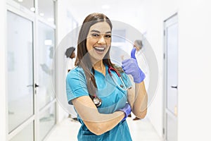 Portrait Of Smiling Female Doctor Wearing Scrubs In Busy Hospital Corridor showing thumbs up