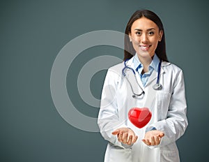 Portrait of smiling female doctor wearing white coat and stethoscope with a heart levitating above her hands and copy space