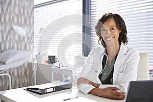 Portrait Of Smiling Female Doctor Wearing White Coat With Stethoscope Sitting Behind Desk In Office