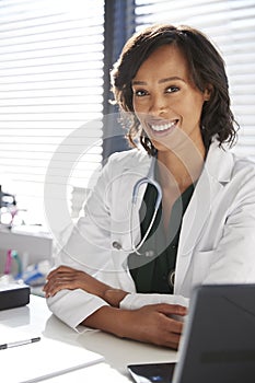 Portrait Of Smiling Female Doctor Wearing White Coat With Stethoscope Sitting Behind Desk In Office