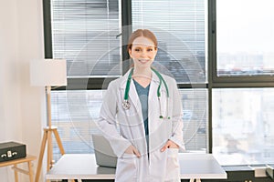 Portrait of smiling female doctor in white coat standing on background of window in sunny day in light medical clinic