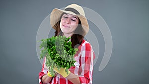 Portrait of smiling female farmer in plaid shirt, gloves and hat holding, smells a bunch of fresh green parsley, greens