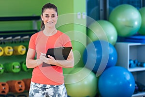 Portrait of smiling female fitness instructor writing in clipboard while standing in gym